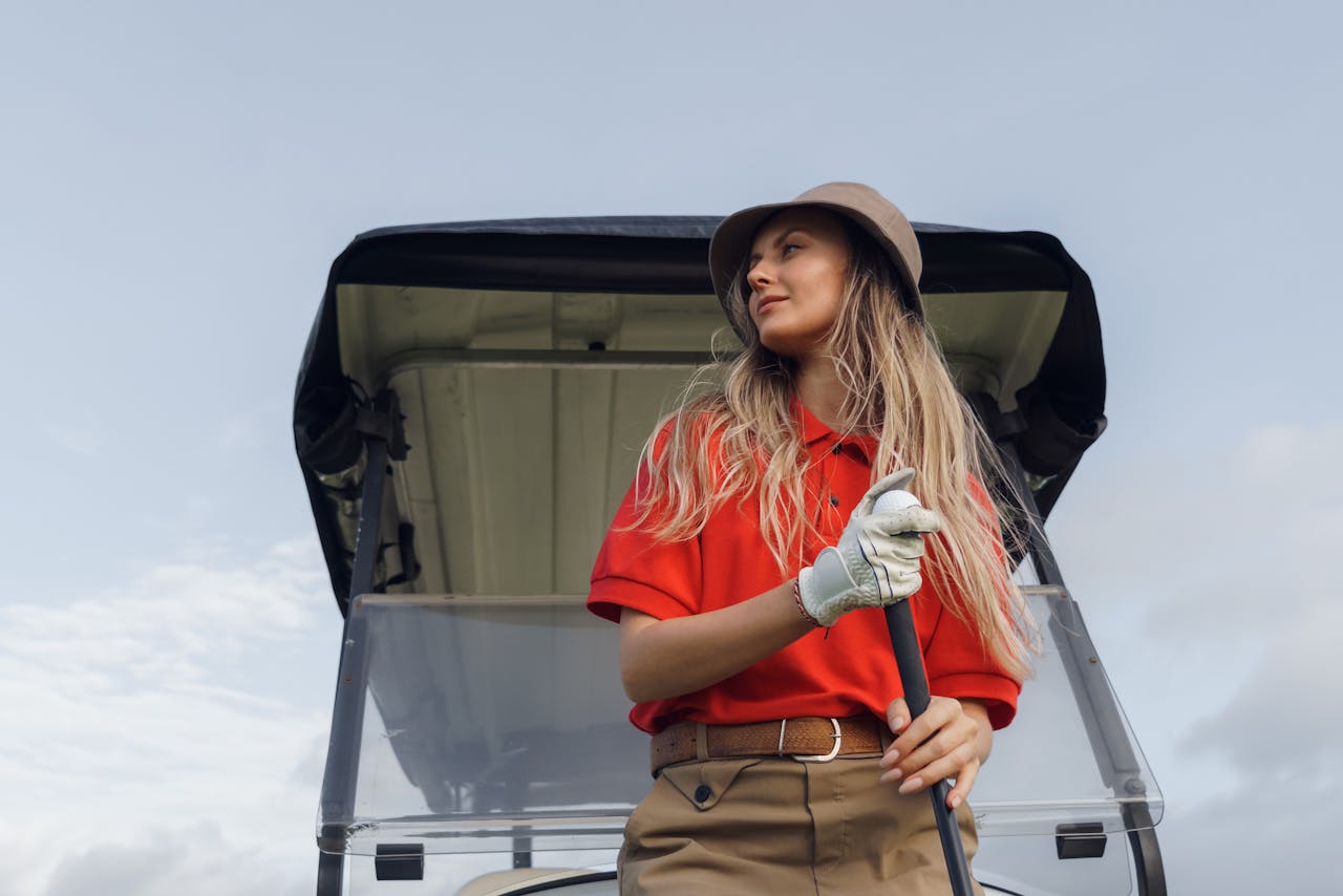 Woman golfer standing by a golf cart, holding a club, wearing red polo, outdoors.